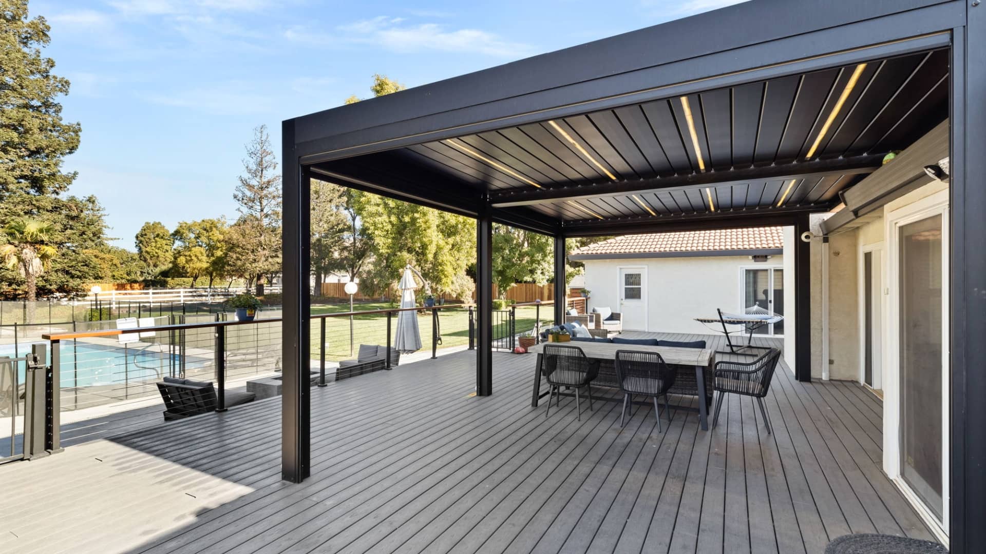 Outdoor living space with a modern black pergola, deck, and patio furniture overlooking a pool and landscaped backyard.