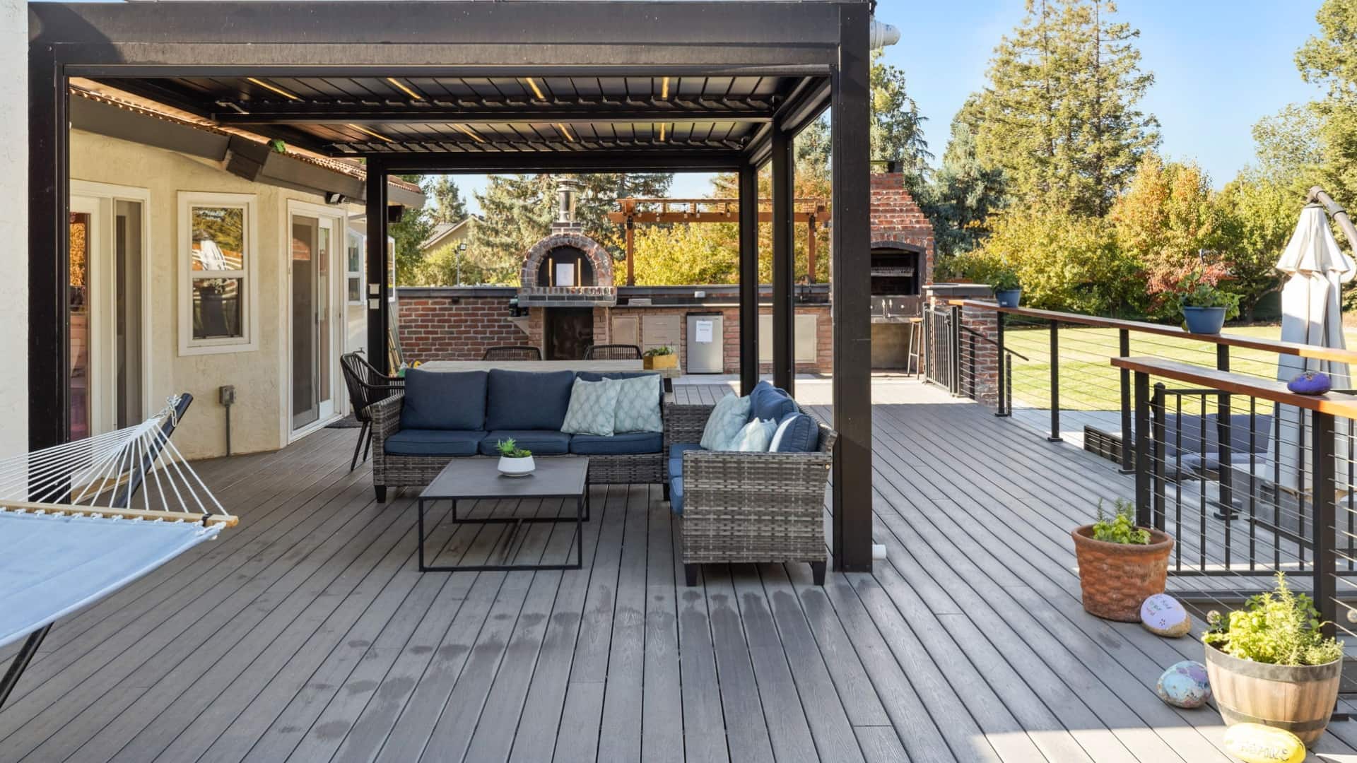 Outdoor deck with pergola, wicker furniture, pizza oven, and grill for backyard relaxation and entertainment.